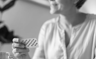 A smiling woman is holding a blister packet or HRT pills and the leaflet, with a drink of water ready on the table sitting at home